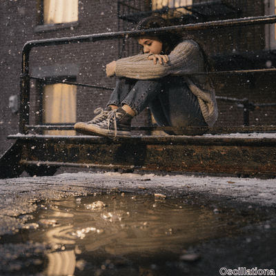 A pensive young woman with long brown hair hugs her knees tightly on a rusty fire escape platform in snowy urban dusk, wearing loose light gray knit sweater, faded jeans rolled at ankles, worn white Converse high-tops (