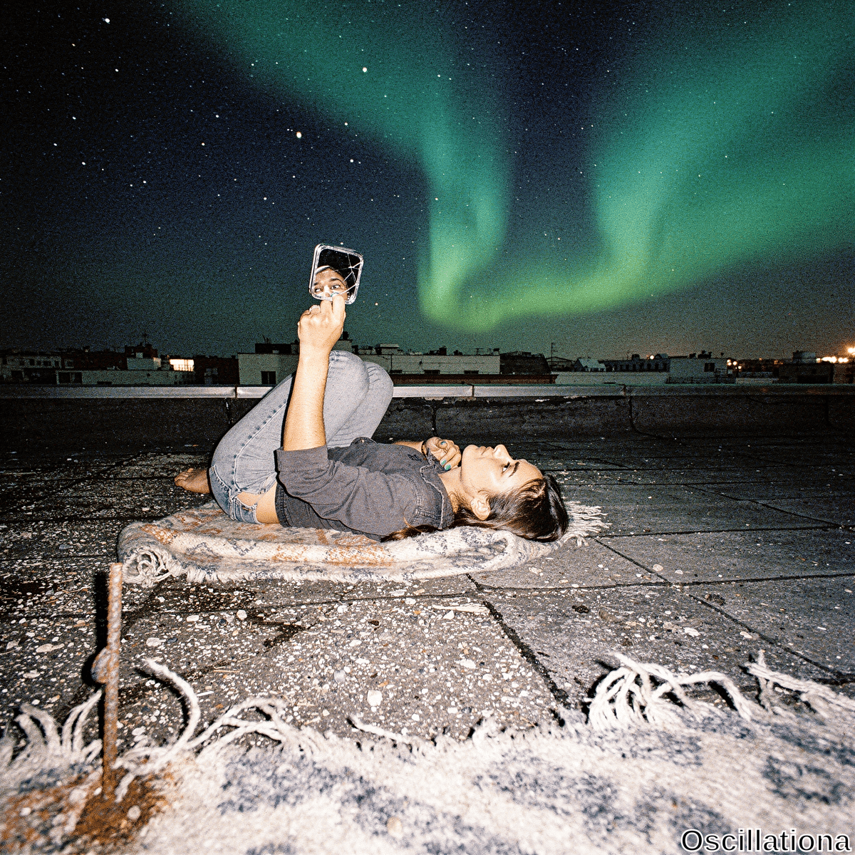 A tan-skinned young woman with dark hair lies relaxed on her back on a gritty urban rooftop at night, holding a cracked silver mirror above her face to capture her upward gaze. Barefoot in ripped light blue jeans and loose gray