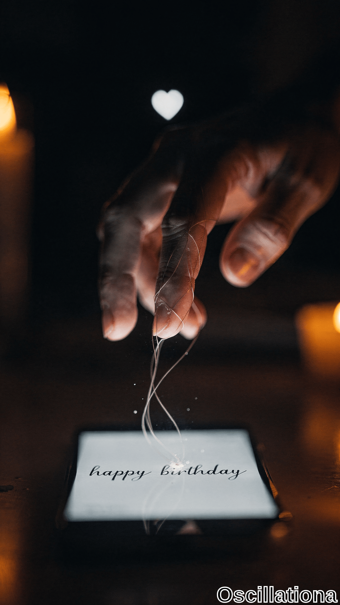 A hand reaches down with delicate glowing threads connecting to a smartphone displaying 'happy birthday' on its screen, set against dark surroundings with warm candlelight and a floating heart. The intimate scene blends realism with surreal, mysterious atmosphere.