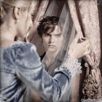 Vintage sepia-toned portrait: blonde woman in profile with loose updo, pearl earring, dusky blue velvet gown & pink lace, parting sheer white veil with pale spread fingers to reveal young man with tousled