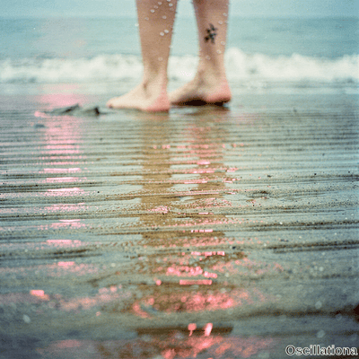 Bare legs and feet with black star tattoo on ankle standing in shallow beach surf as waves ripple reflecting pink golden light in dreamy film style.
.