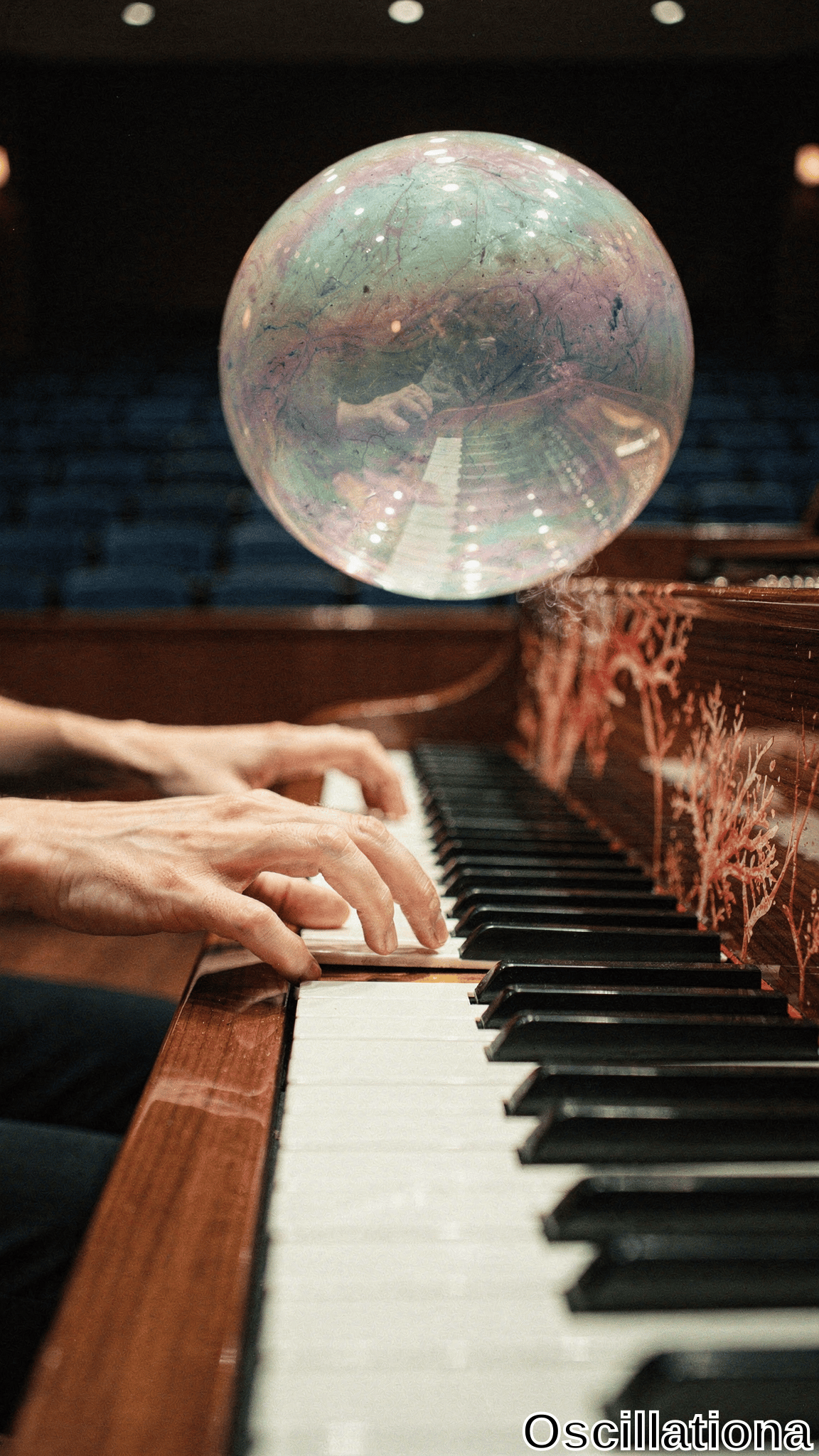 Surreal close-up in dimly lit auditorium: large iridescent soap bubble floats above grand piano, shimmering teal, purple, pink, green hues reflecting blurred veined hands on keys, blue seats. Reddish-brown piano with