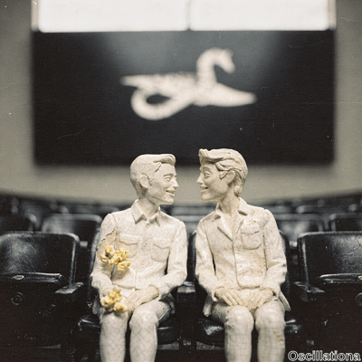 Sepia-toned diorama of two cracked porcelain figurines of young men on black leather theater seats, gazing affectionately in dim cinema light. Left with dark hair holds yellow flowers, smiling; right blondish-haired smiles gently;