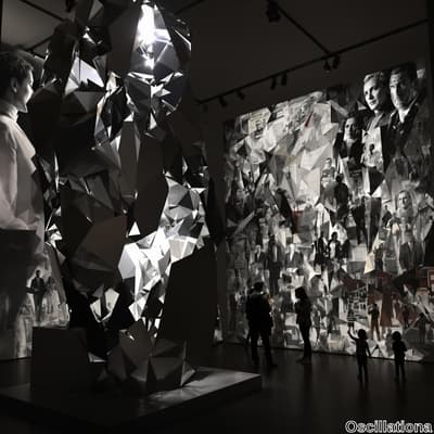 A large, abstract sculpture with reflective, angular facets stands in a dimly lit gallery, casting bright highlights.